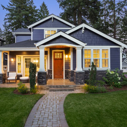 facade of home with manicured lawn, landscaping, and backdrop of trees and dark blue sky. Glowing interior lights create a welcoming mood.